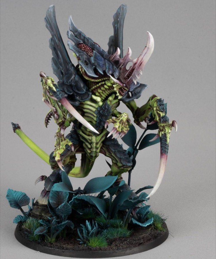 A large insectoid alien miniature painted sickly yellow-green with dark slate-blue armor plates and pale bone talons, on a base of teal plants.