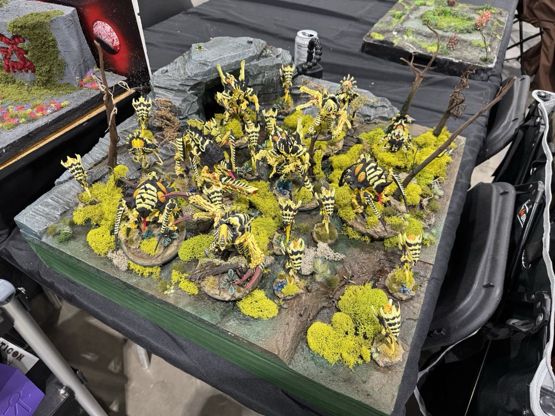 A whole army of alien miniatures painted in bright yellow with black stripes, displayed on a mossy display board at a convention.